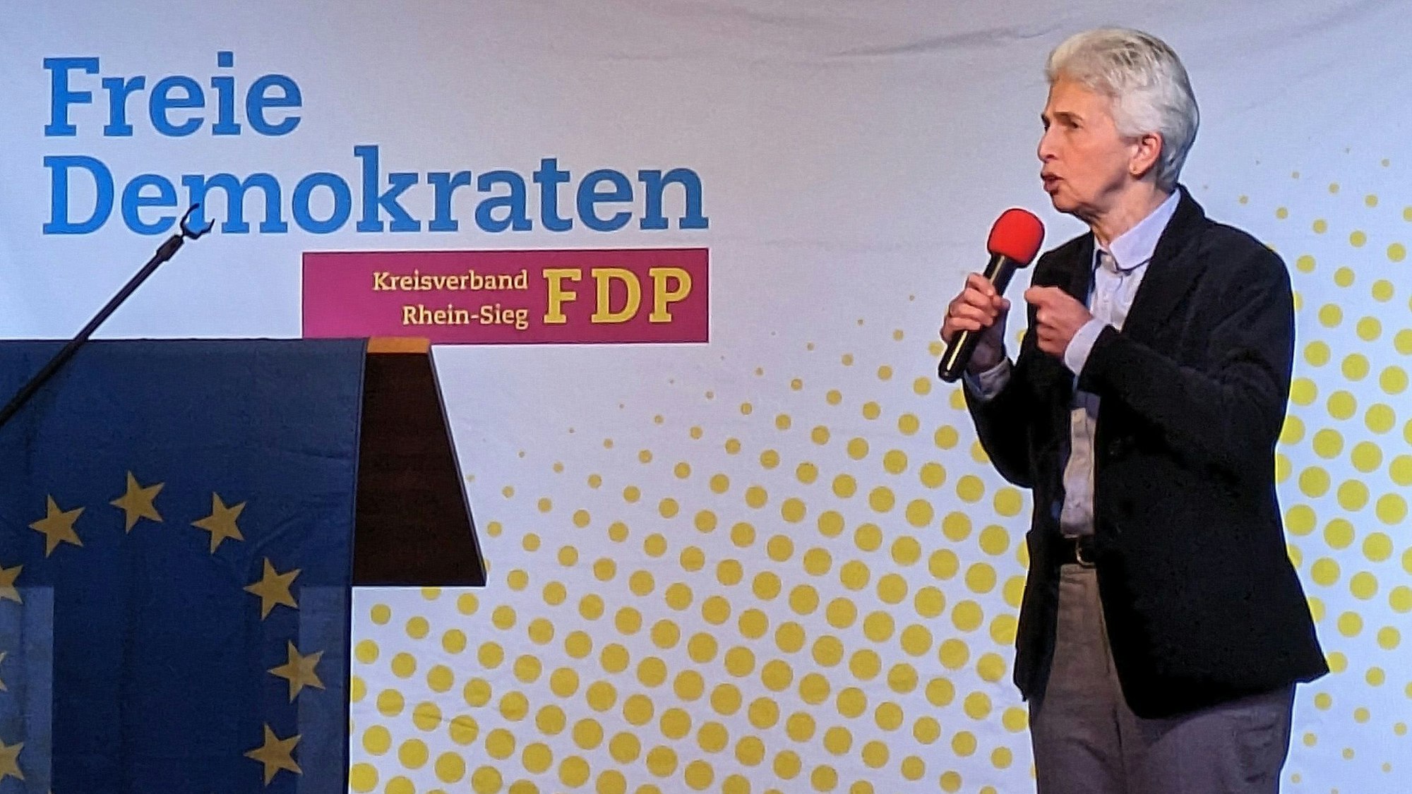 Eine Frau mit kurzen grauen Haaren, schwarzem Sakko und grauer Hose spricht in ein Mikrofon. Auf der Wand steht Freie Demokraten.
