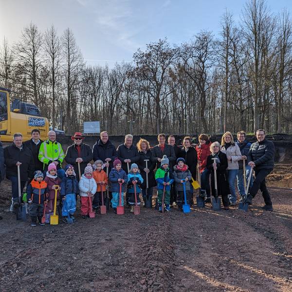 Eine Gruppe von Kindern mit Schaufeln und Erwachsenen mit Spaten auf einer Baustelle.