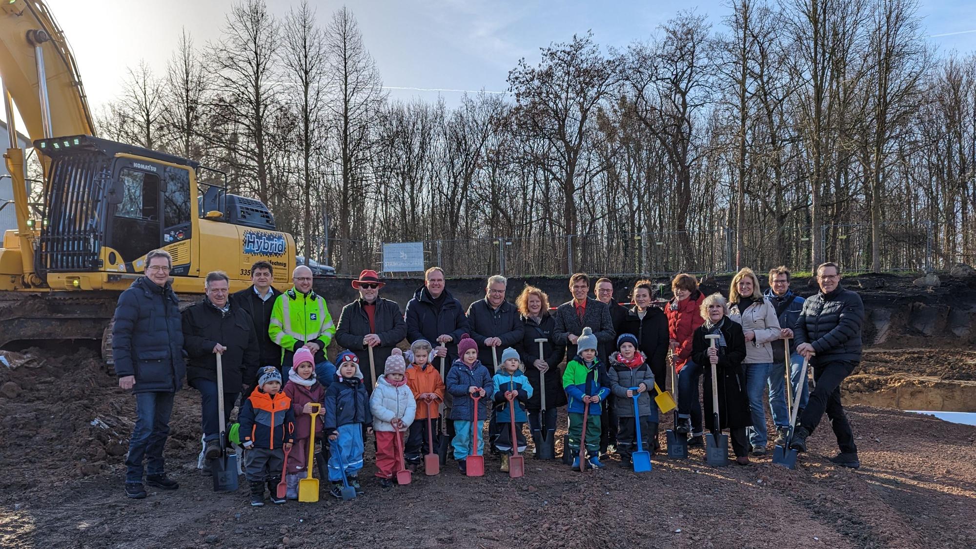 Eine Gruppe von Kindern mit Schaufeln und Erwachsenen mit Spaten auf einer Baustelle.