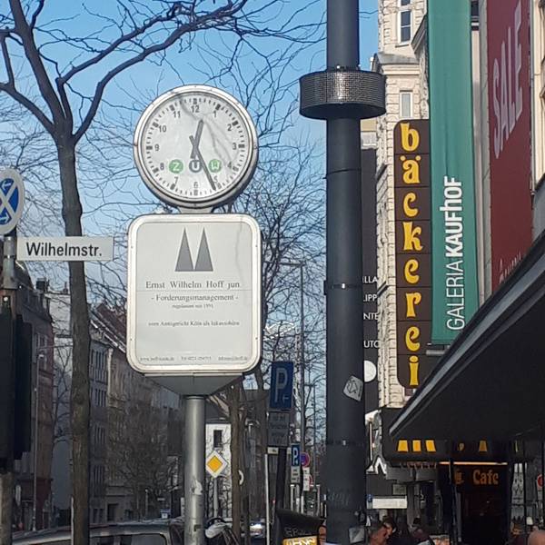 An einem Laternenmast ist ein rundes Display angebracht, daneben ist eine Uhr, ein Straßenschild und das Gebäude von Galeria Kaufhof zu sehen.
