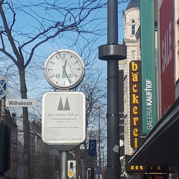 An einem Laternenmast ist ein rundes Display angebracht, daneben ist eine Uhr, ein Straßenschild und das Gebäude von Galeria Kaufhof zu sehen.