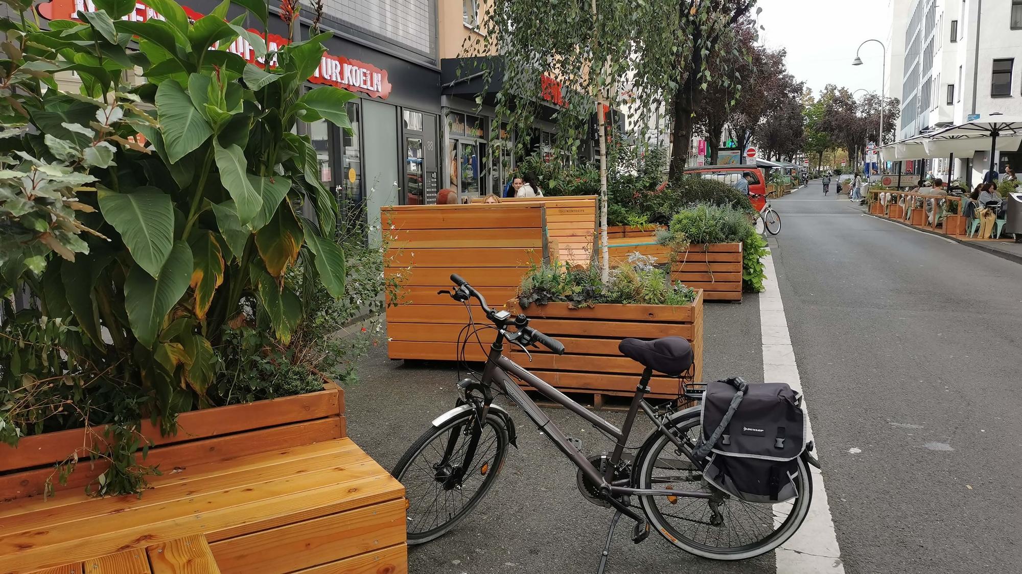 Auf einem Parkstreifen stehen Holzkübel mit Blume und Sitzmöglichkeiten.