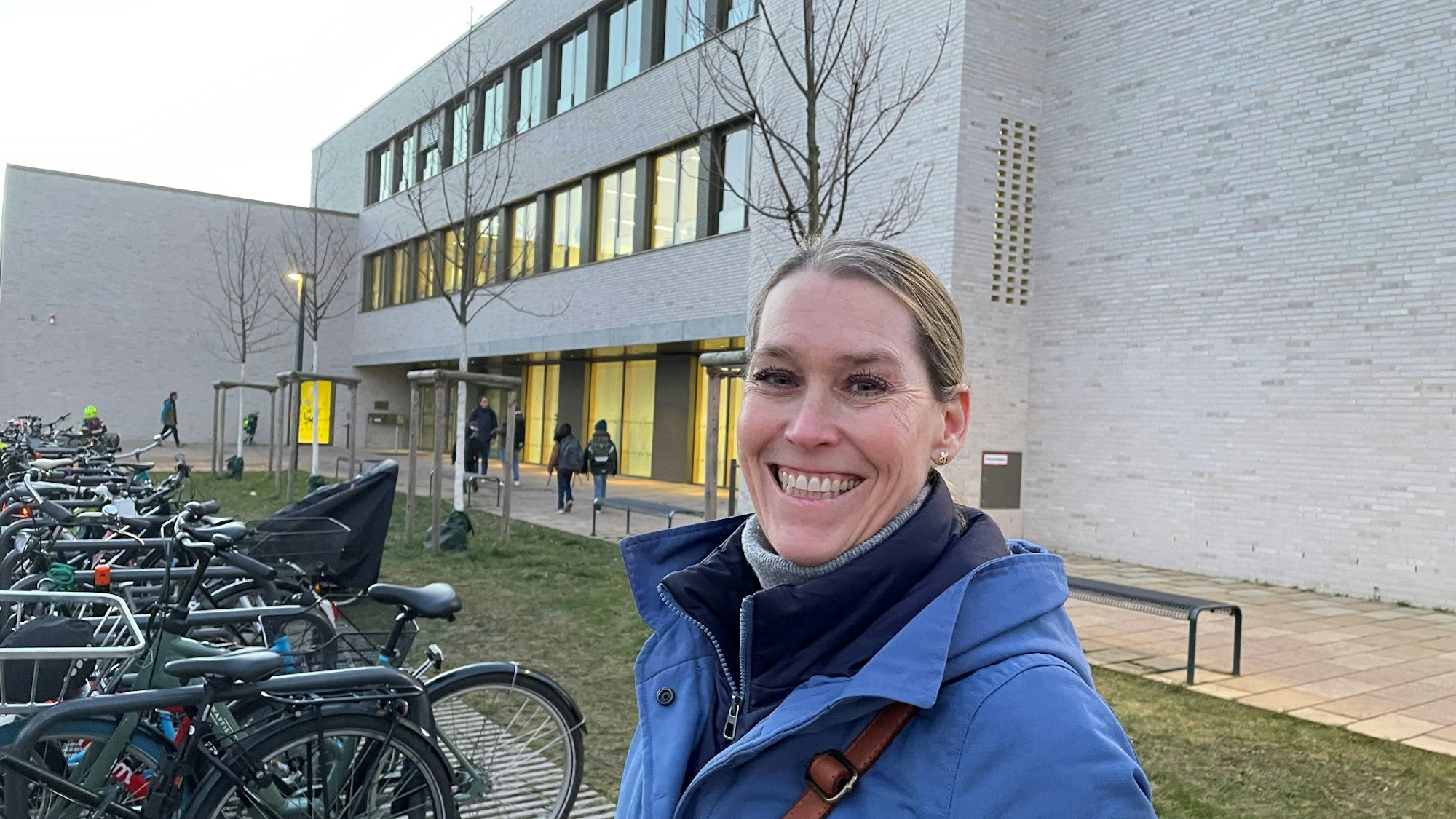 Eine Frau mit Zopf steht lachend vor einem Schulgebäude.