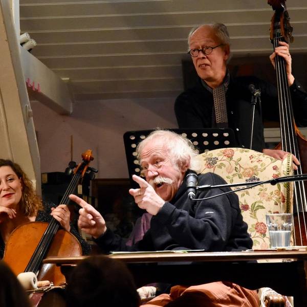 Ein Mann mit weißen Haaren und Schnurrbart rezitiert; er sitzt dabei auf einem Sessel. Eine Frau mit Cello sitzt links, ein Mann mit Kontrabass steht im Hintergrund.