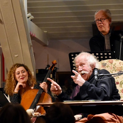 Ein Mann mit weißen Haaren und Schnurrbart rezitiert; er sitzt dabei auf einem Sessel. Eine Frau mit Cello sitzt links, ein Mann mit Kontrabass steht im Hintergrund.