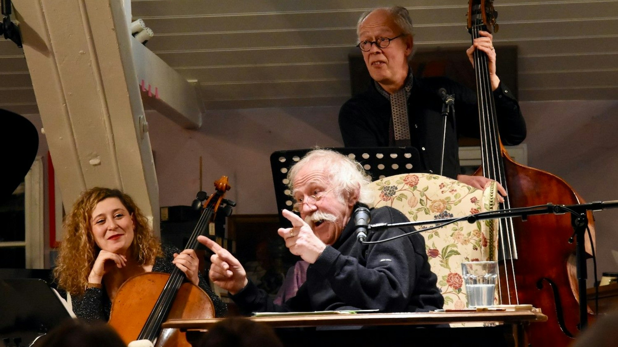 Ein Mann mit weißen Haaren und Schnurrbart rezitiert; er sitzt dabei auf einem Sessel. Eine Frau mit Cello sitzt links, ein Mann mit Kontrabass steht im Hintergrund.