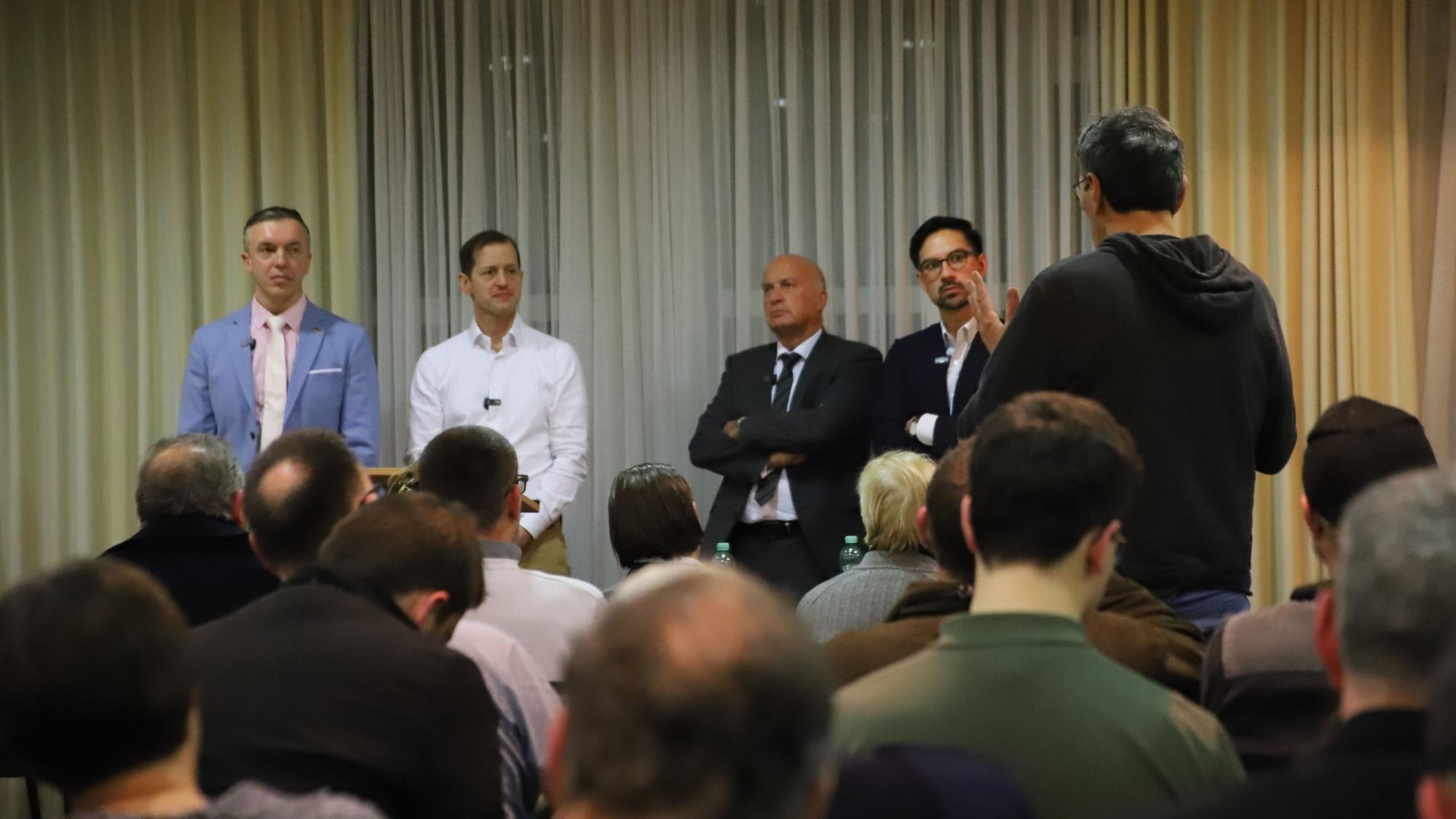 Männer in Anzügen stehen vor Publikum..