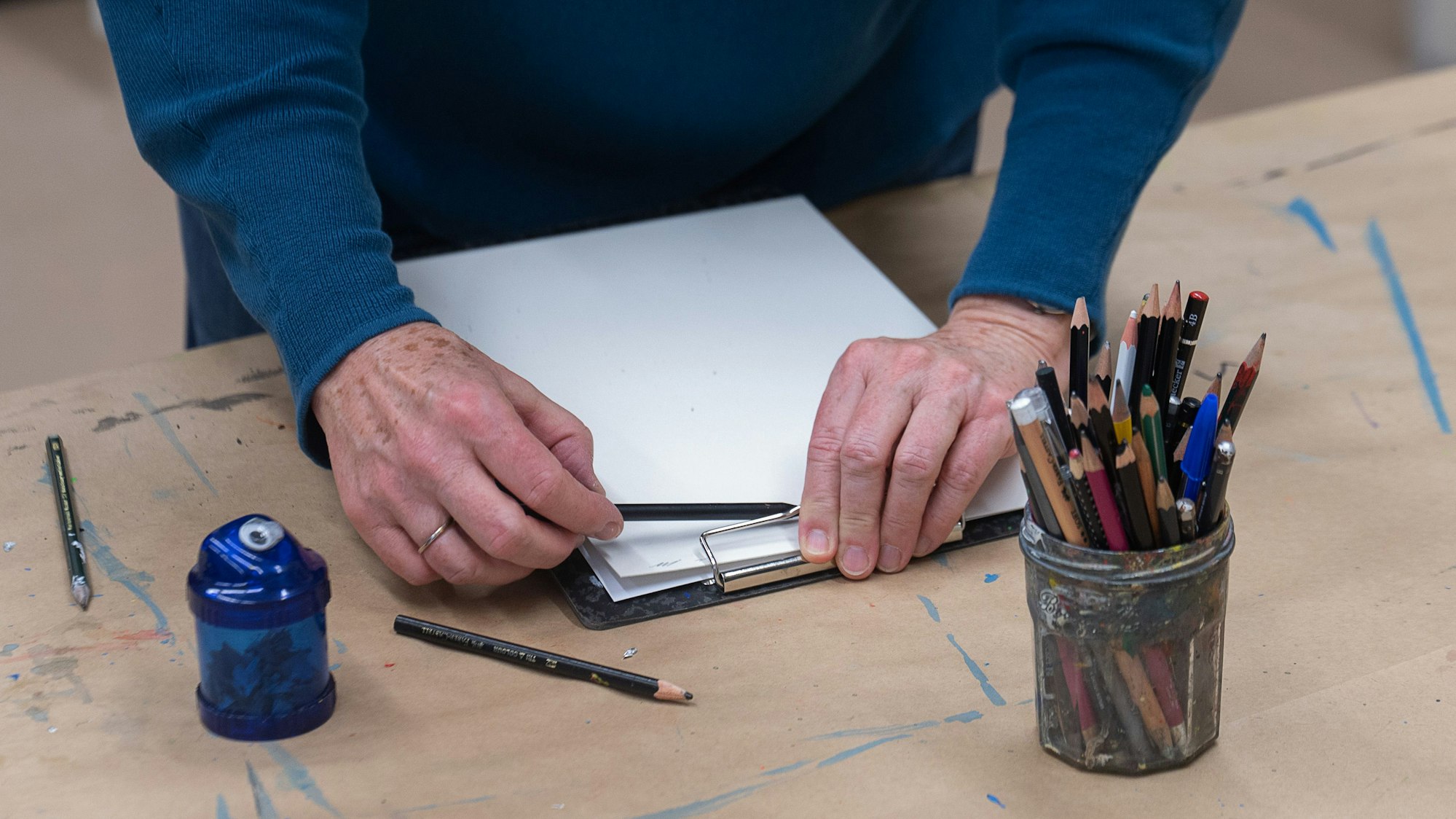 Ein Mann hält einen Stift. Vor ihm liegt ein Klemmbrett mit weißen leeren Blättern. Vor ihm steht ein Glas mit mehreren Stiften sowie ein blauer Anspitzer.