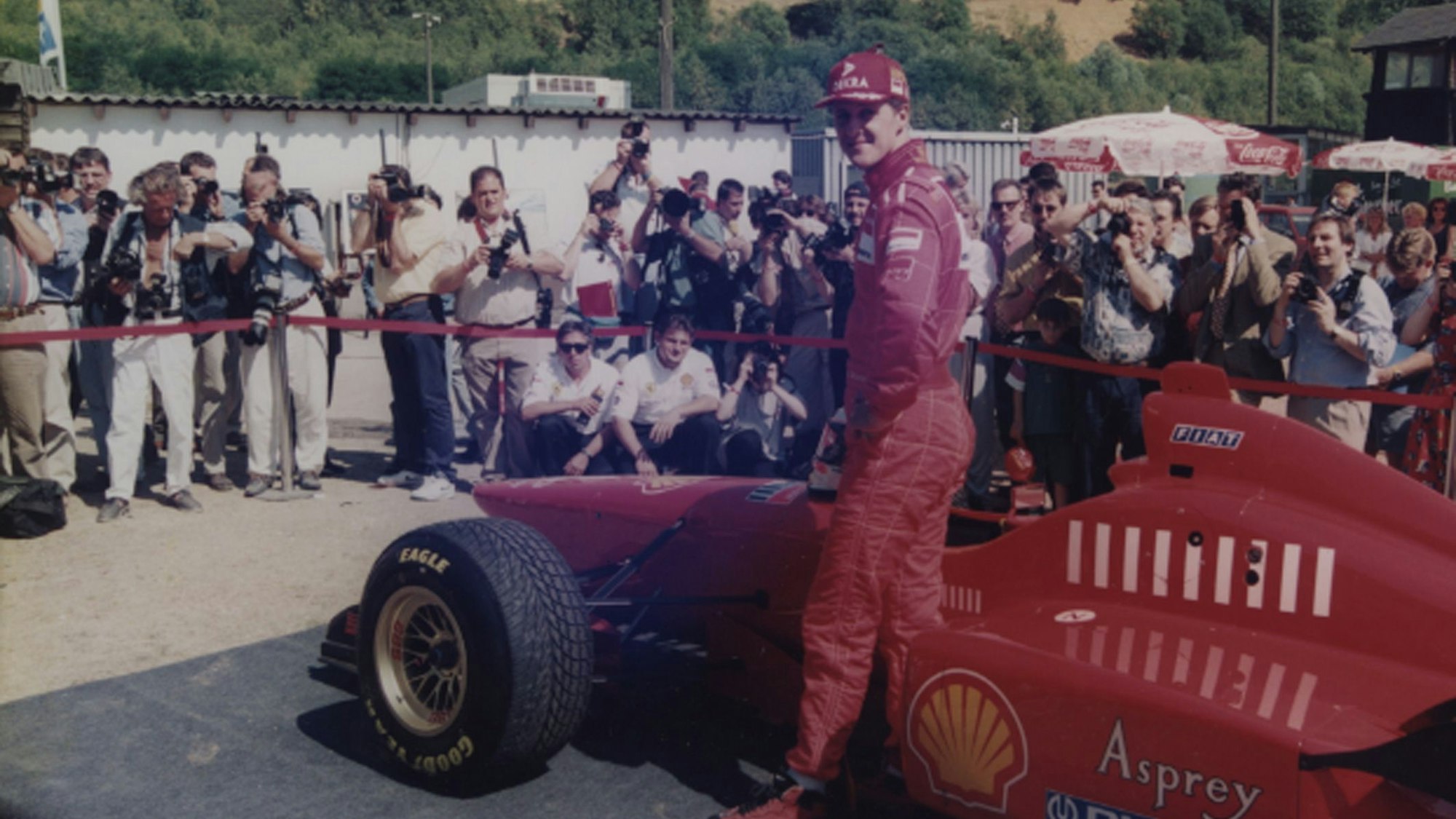 Auf dem Foto ist der Rennfahrer Michael Schumacher vor einem roten Rennwagen zu sehen.