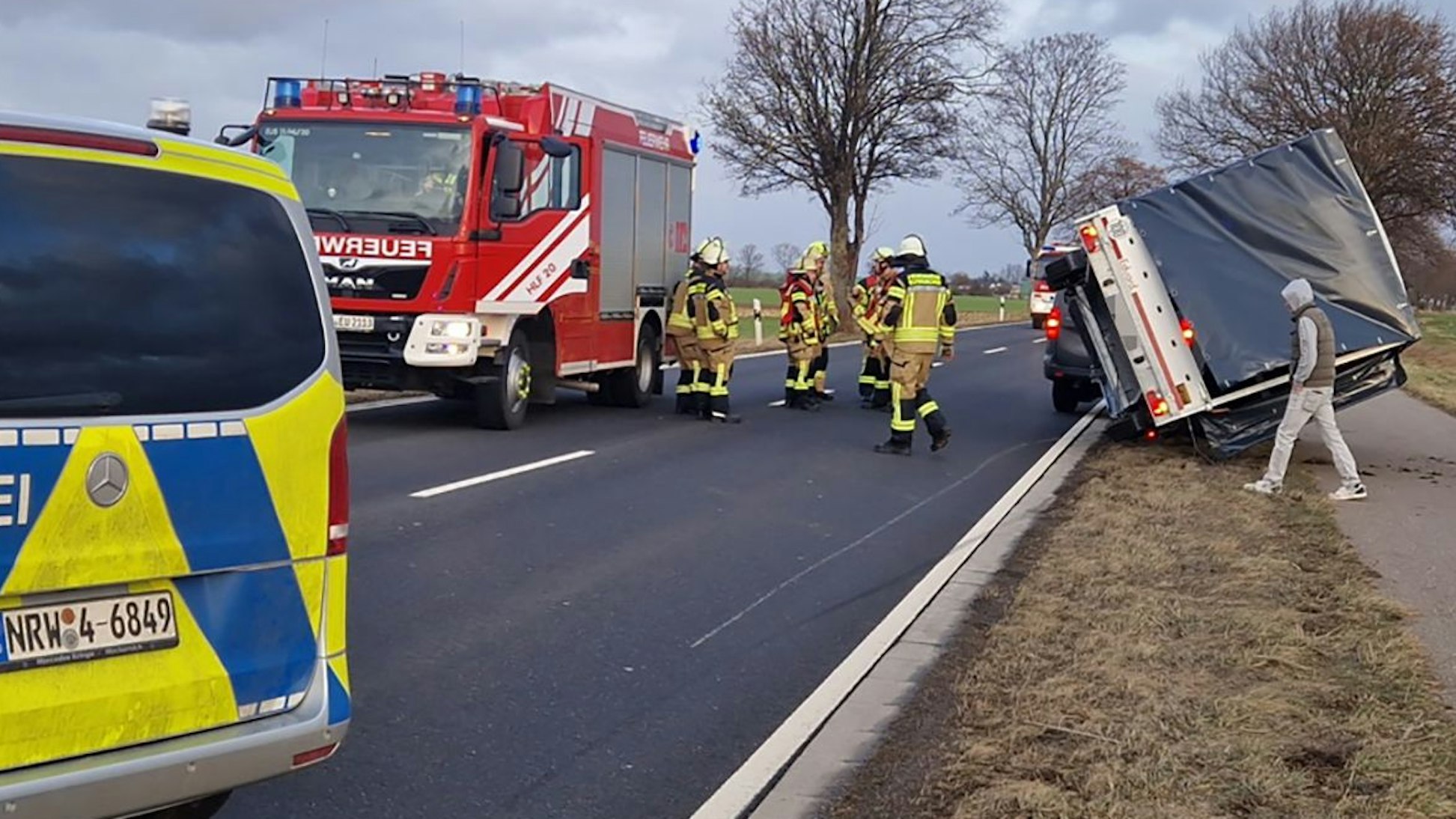 Ein Lkw-Anhänger liegt am Rande der Straße, ein Feuerwehrwagen steht daneben, dazwischen beraten Feuerwehrleute die Lage. Vorne steht ein Polizeiwagen, um die Straße abzusperren.
