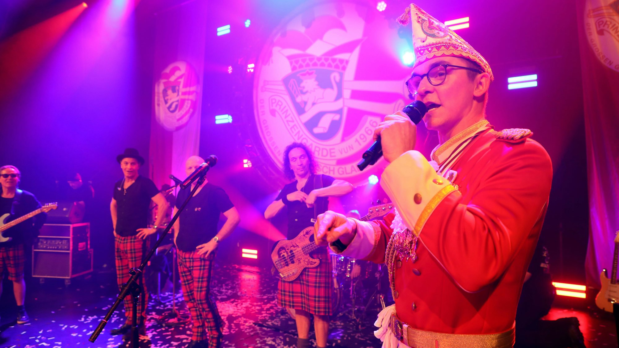 Prinzengarde-Kommandant Marco Wasserberg spricht in ein Mikrofon, im Hintergrund sind Musiker von Brings zu sehen.