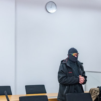Ein Mann in schwarzer Jacke, Sturmhaube und verspiegelter Sonnenbrille steht mit gefalteten Händen in einem Gerichtssaal, an der Wand hängt ein Kreuz.