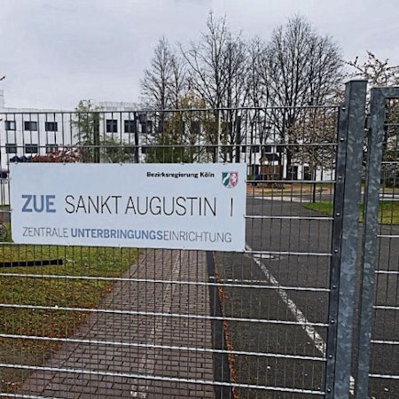 Ein Schild an einem Zaun weist auf dem Gelände an der Heerstraße in Sankt Augustin auf die Zentrale Unterbringungseinrichtung (ZUE) hin.