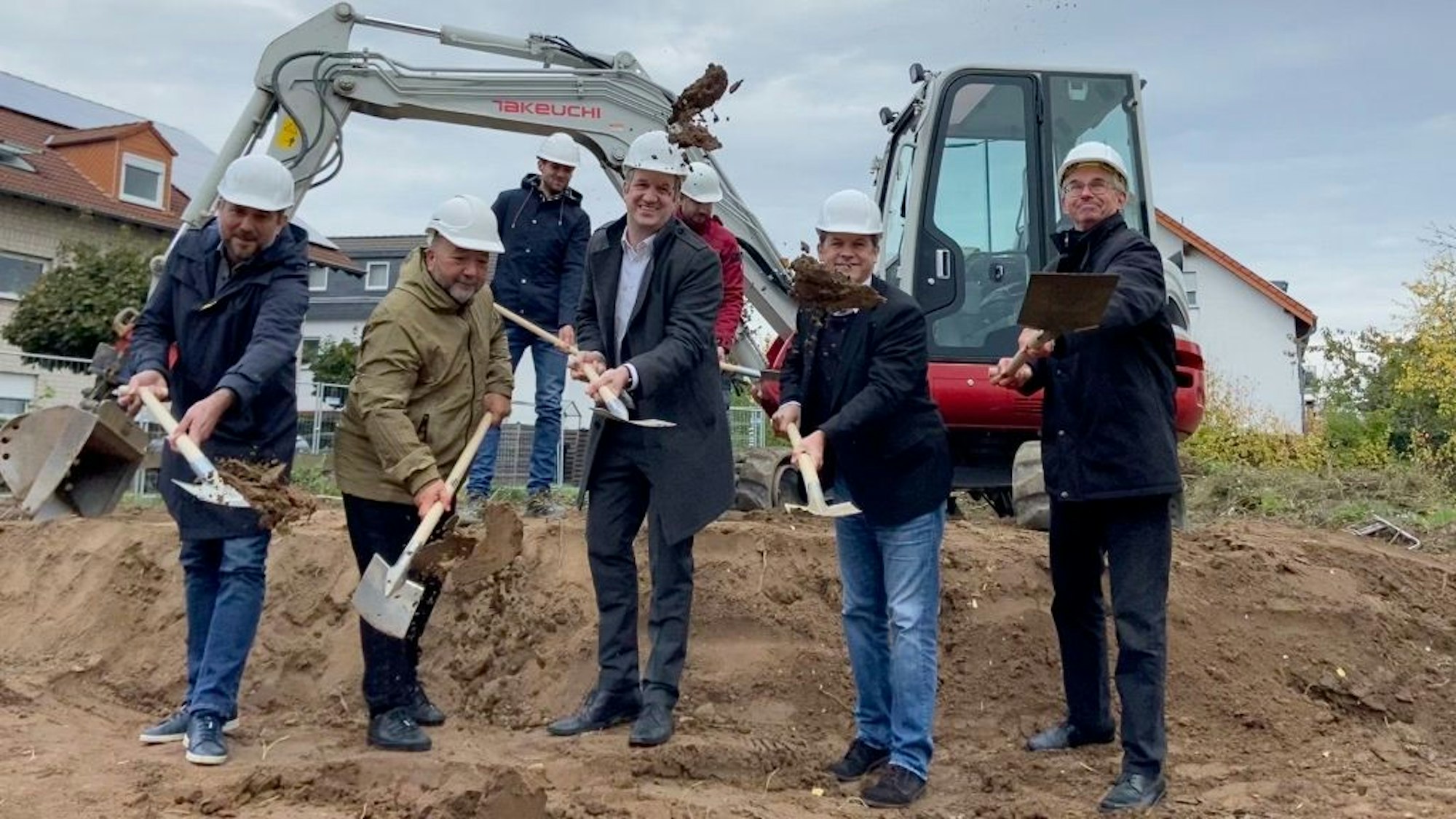 Fünf Männer stehen auf einem Sandhaufen, sie schaufeln Sand, im Hintergrund steht ein Bagger.