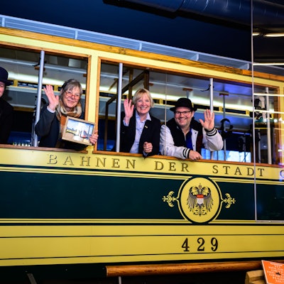 Vier Personen gucken und winken aus einer alten Straßenbahn.