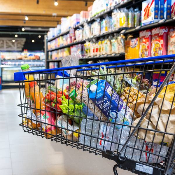Ein voller Einkaufswagen biegt in einen Gang im Supermarkt zwischen zwei Regalen.