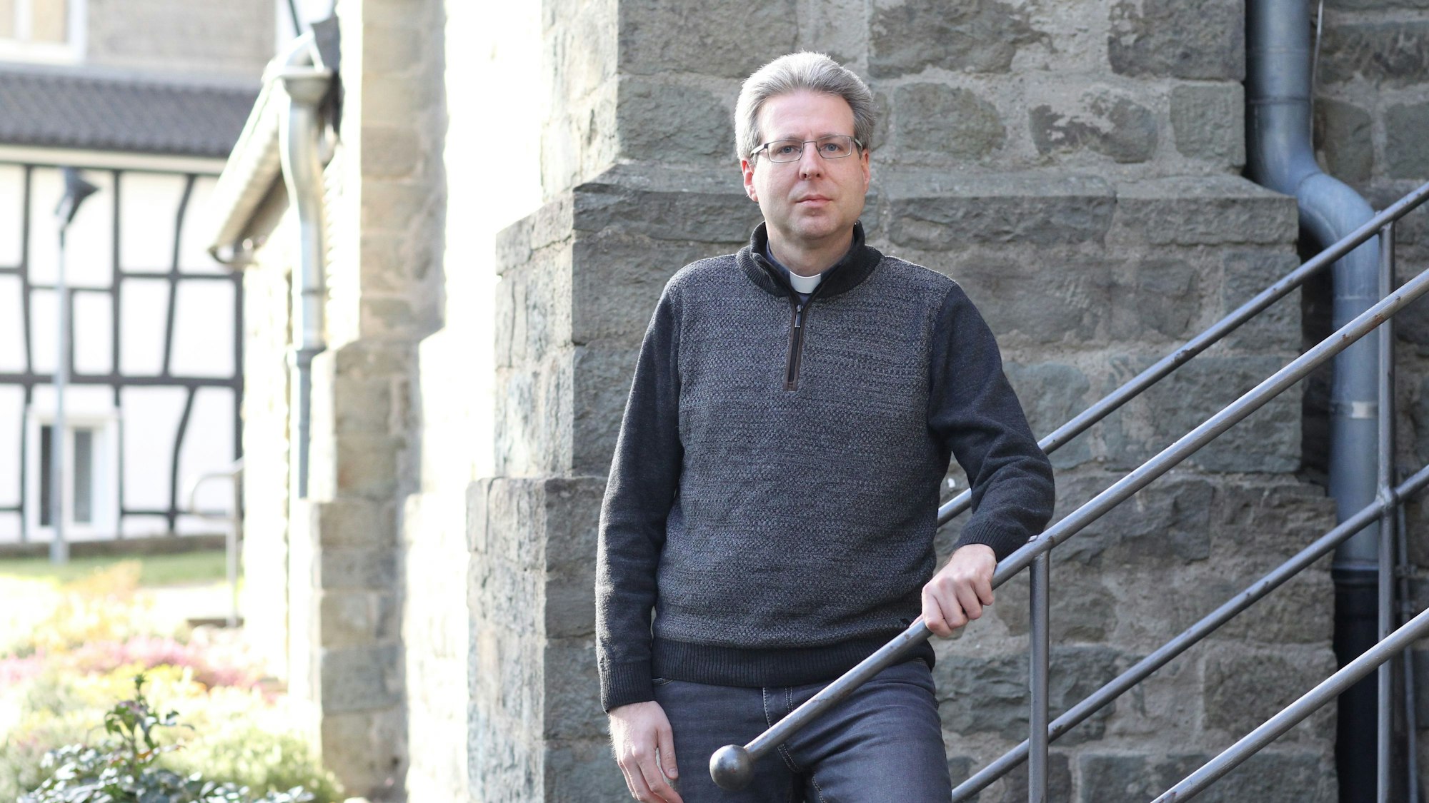 Ein grauhaariger Pfarrer steht auf einer Treppe an einem Kirchengebäude.