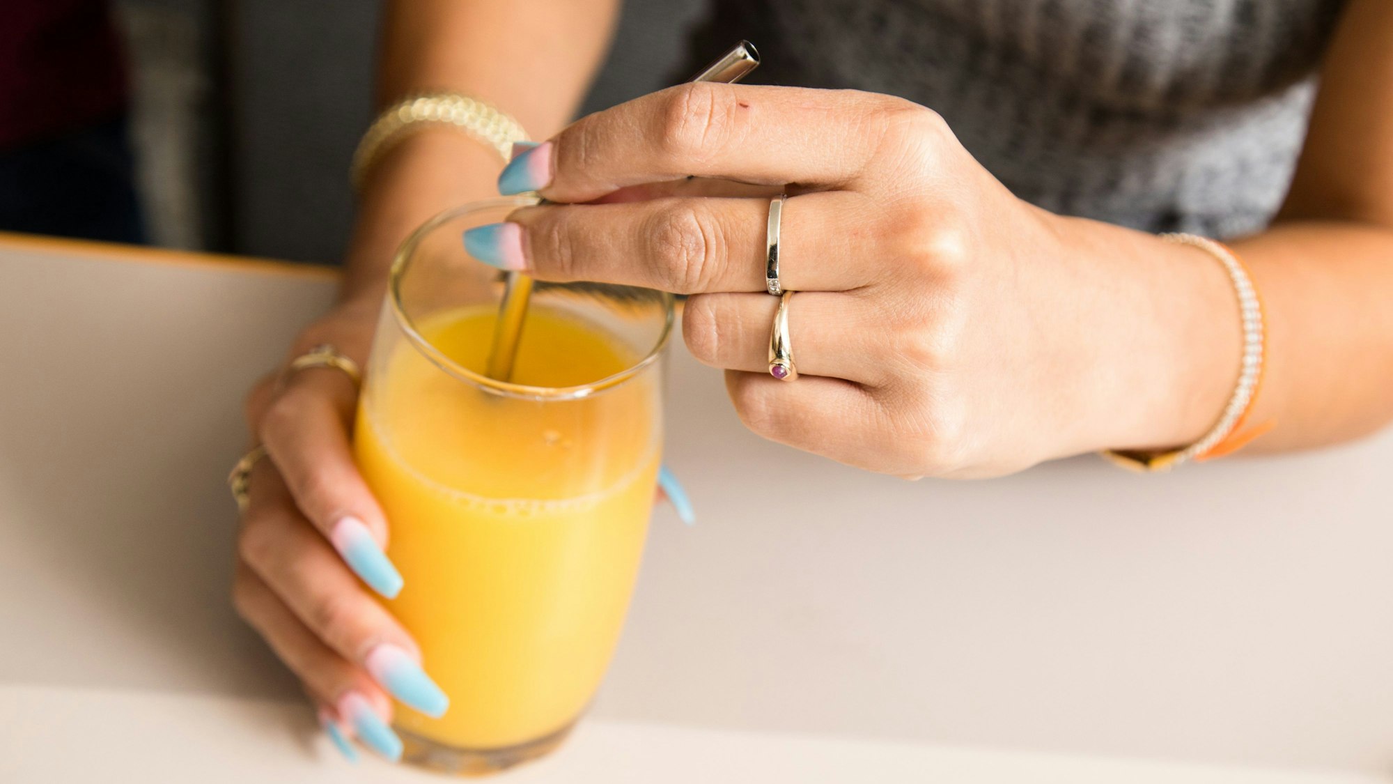 Eine Frau mit bunten Fingernägeln trinkt mit einem Strohhalm aus einem Glas Orangensaft.