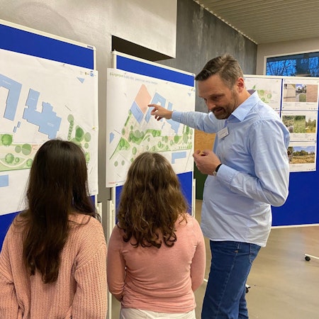 Zu sehen ist ein Mitarbeiter des Planungsbüros, der zwei Kindern die Skizzen des Parks erläutert.