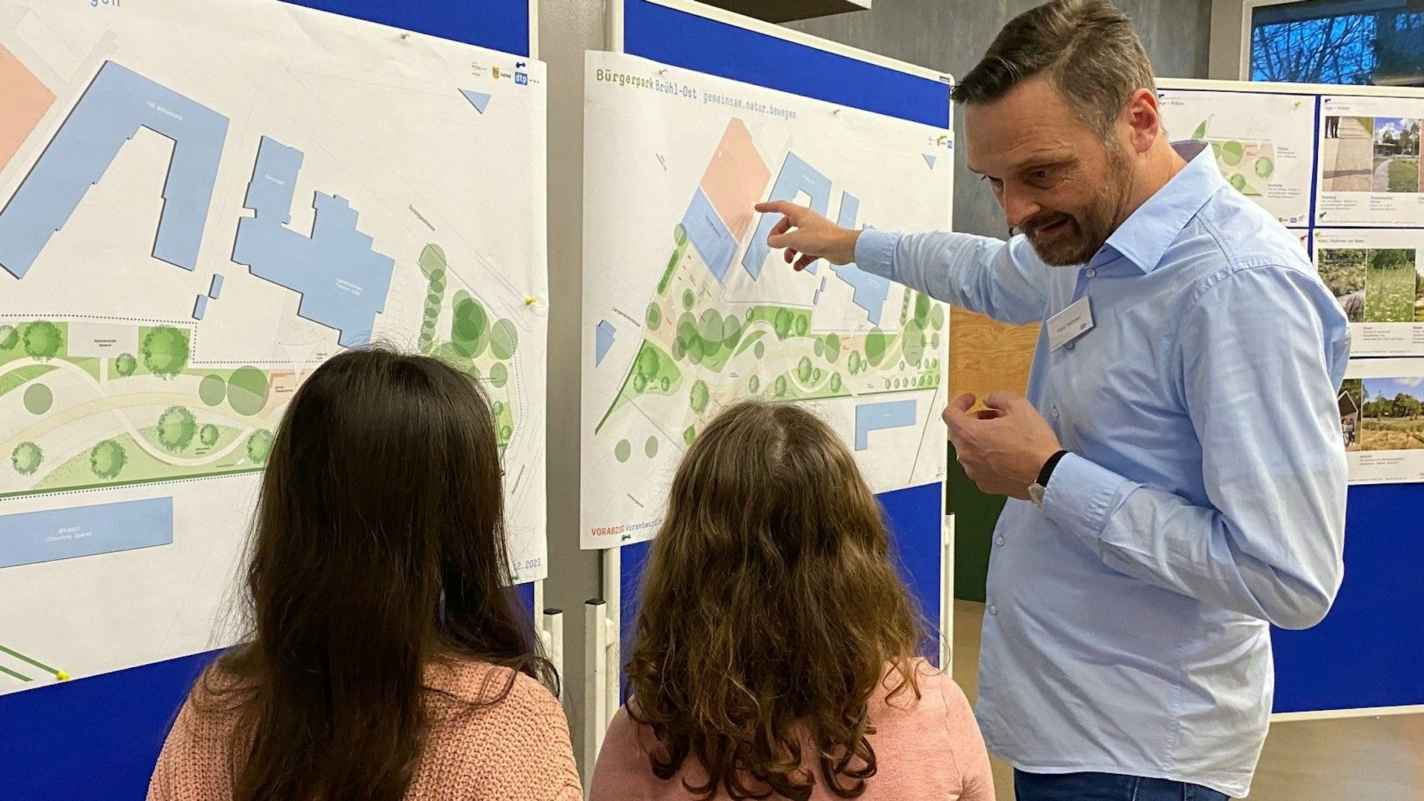 Zu sehen ist ein Mitarbeiter des Planungsbüros, der zwei Kindern die Skizzen des Parks erläutert.