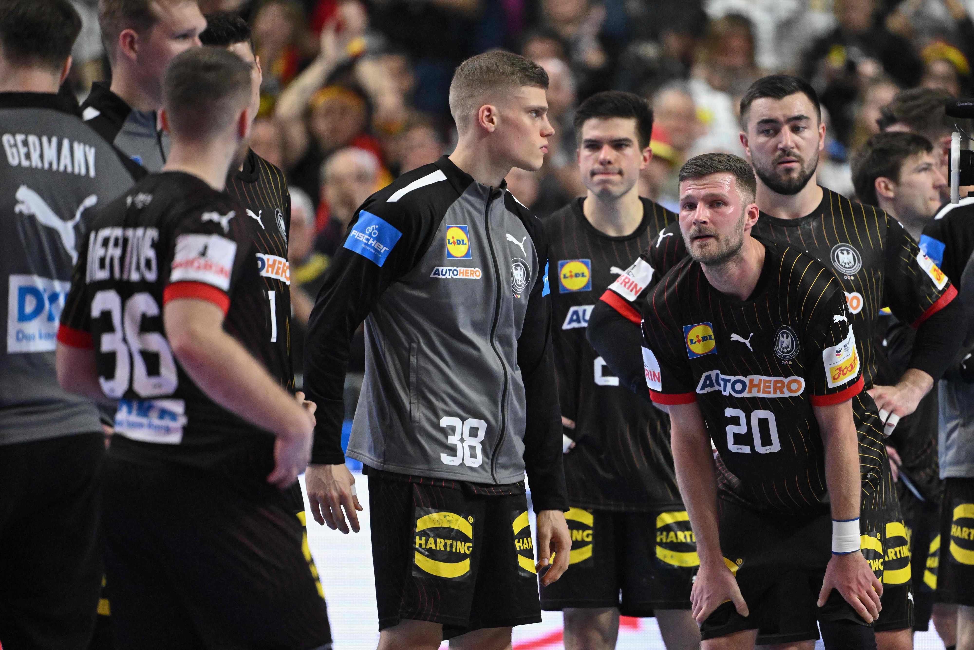 Mehrere Handballspieler stehen erschöpft und niedergeschlagen auf dem Spielfeld. Im Hintergrund befinden sich verschwommen aufgenommene Zuschauende.