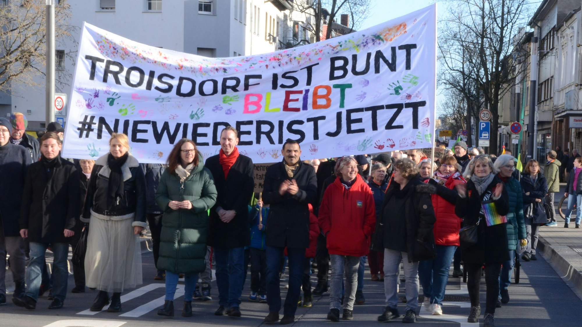 Ein Protestzug, in der ersten Reihe gehen Männer und Frauen unter einem Transparent. Darauf steht "Troisdorf ist bunt" und "Nie wieder ist jetzt"