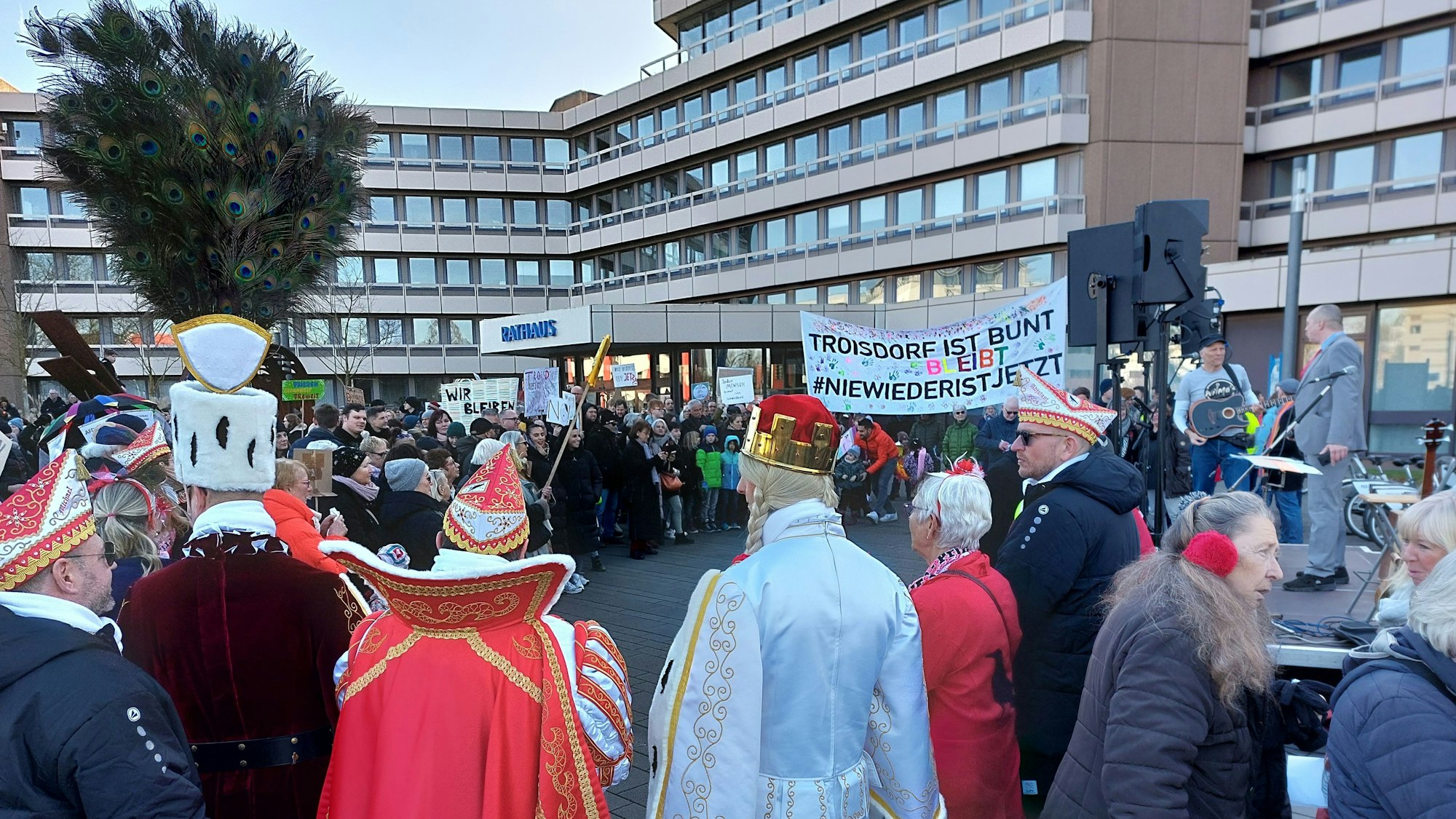 Vor einem großen Verwaltungsgebäude stehen viele Menschen. Sie halten Transparente in die Höhe. Im Vordergrund steht ein Karnevalsdreigestirn: Prinz, Bauer und Jungfrau.