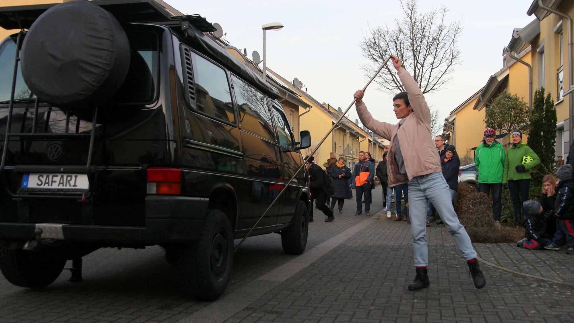 Mit einem Wagenheber hebt die Künstlerin Tanja Kodlin einen schwarzen Wagen an. Anwohner beobachten die Performance.