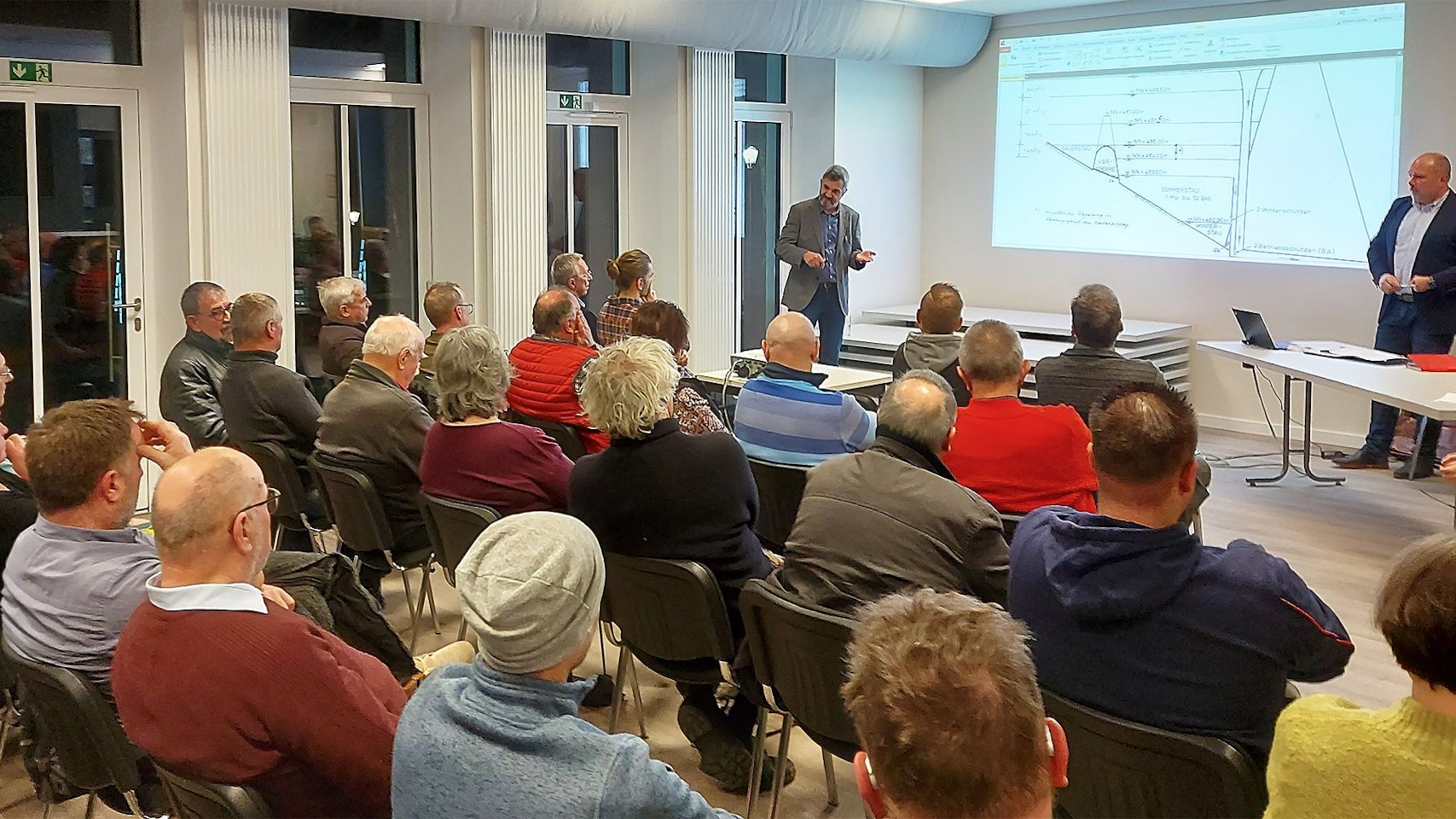 Menschen sitzen in einem Raum und hören einem Vortrag zu, der per Beamer an eine Wand gestrahlt wird.