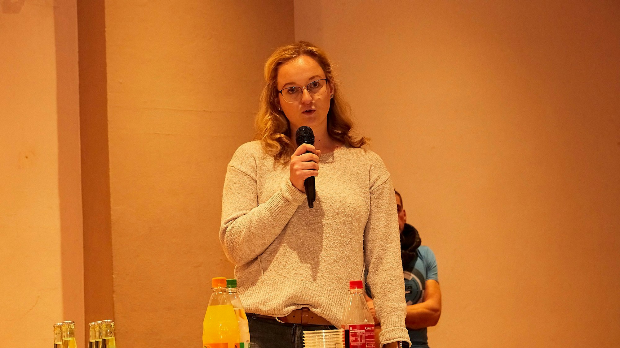 Janina Weins steht mit einem Mikrofon in der Hand an einem Tisch, auf dem Softdrinks zu sehen sind.