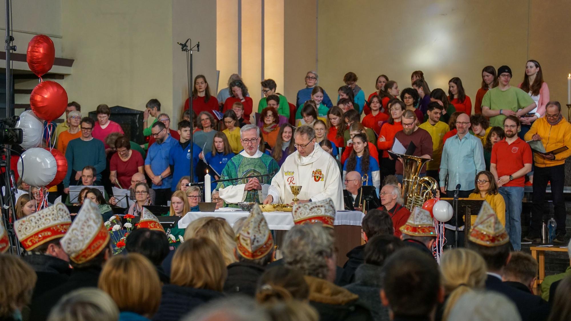 Blick von den voll besetzten Kirchenbänken zum Altar, wo ein Pfarrer und ein Diakon stehen, dahinter ein Chor, bestehend aus circa 50 bunt angezogenen Männern und Frauen.