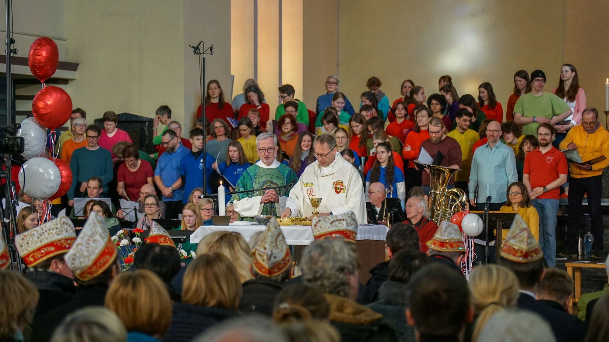 Blick von den voll besetzten Kirchenbänken zum Altar, wo ein Pfarrer und ein Diakon stehen, dahinter ein Chor, bestehend aus circa 50 bunt angezogenen Männern und Frauen.