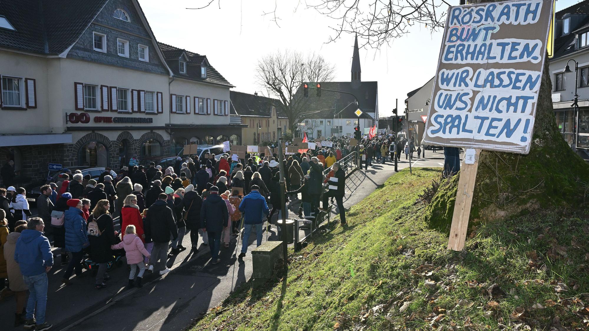 Ein Schild mit der Aufschrift „Rösrath bunter erhalten – wir lassen uns nicht spalten“ lehnt an einem Baum. Auf der Straße ist ein Demonstrationszug zu sehen.