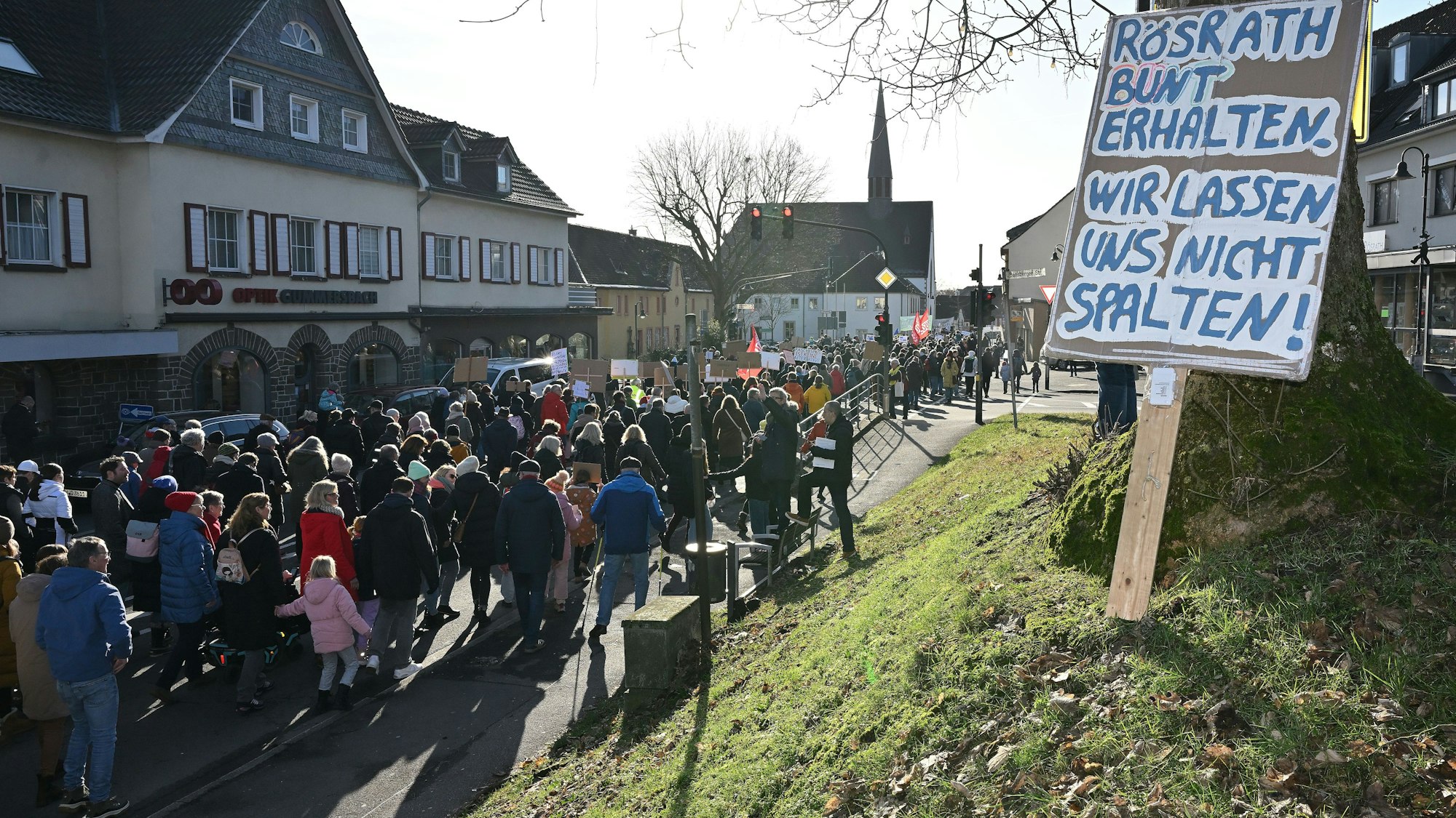Ein Schild mit der Aufschrift „Rösrath bunter erhalten – wir lassen uns nicht spalten“ lehnt an einem Baum. Auf der Straße ist ein Demonstrationszug zu sehen.