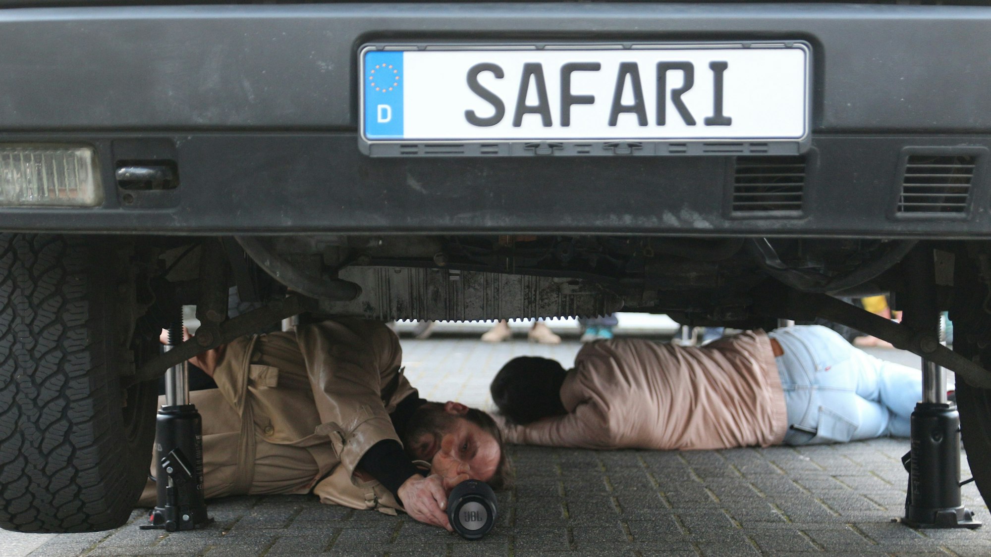 Auf dem Foto sind zwei Künstler zu sehen, die auf der Straße unter einem Fahrzeug liegen.