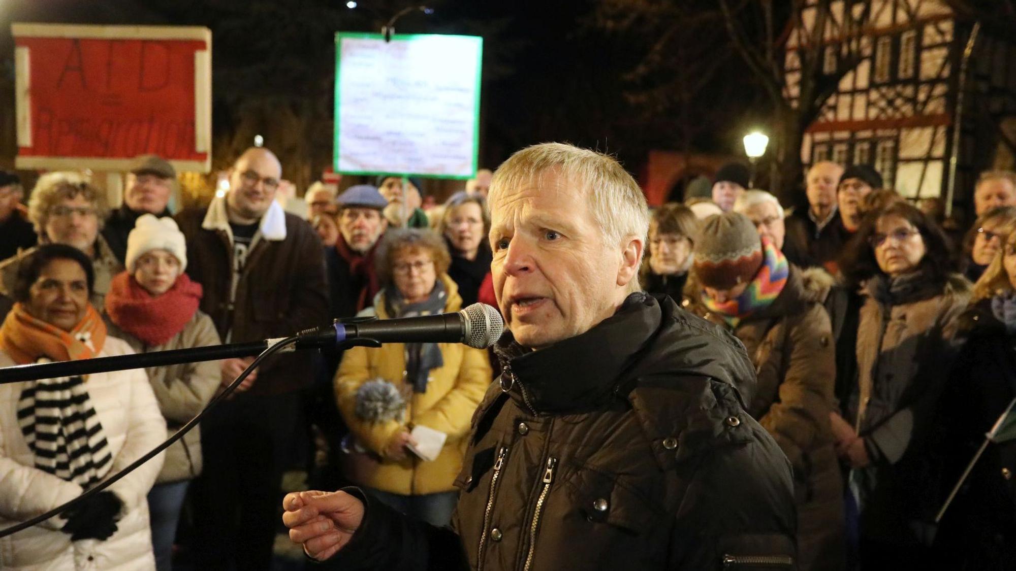Ein Mann in schwarzer Winterjacke spricht zu Teilnehmern einer Versammlung auf einem öffentlichen Platz.