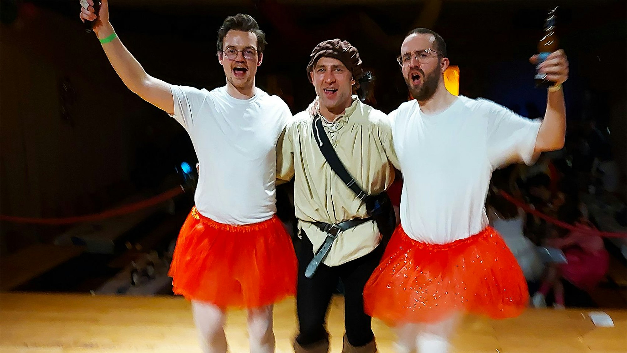 Tobis Schiffer und Andreas Blindert mit DJK-Präsident Salih Sahim in der Mitte stehen bunt kostümiert und mit Bierflaschen in der Hand feiernd auf einer Bühne.