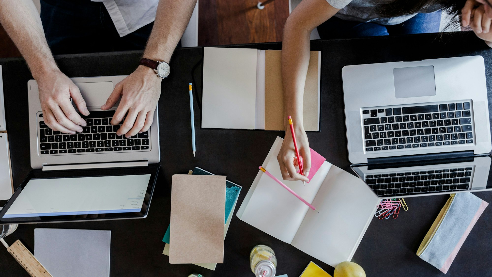 zwei Menschen am Schreibtisch mit Laptops, Notizbüchern und Zetteln