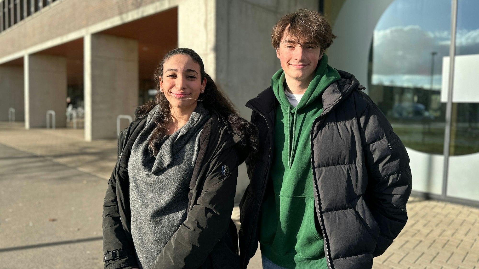 Ein Mädchen mit dunklem Haaren und Pferdeschwanz und ein junger Mann stehen nebeneinander vor einem Gebäude.