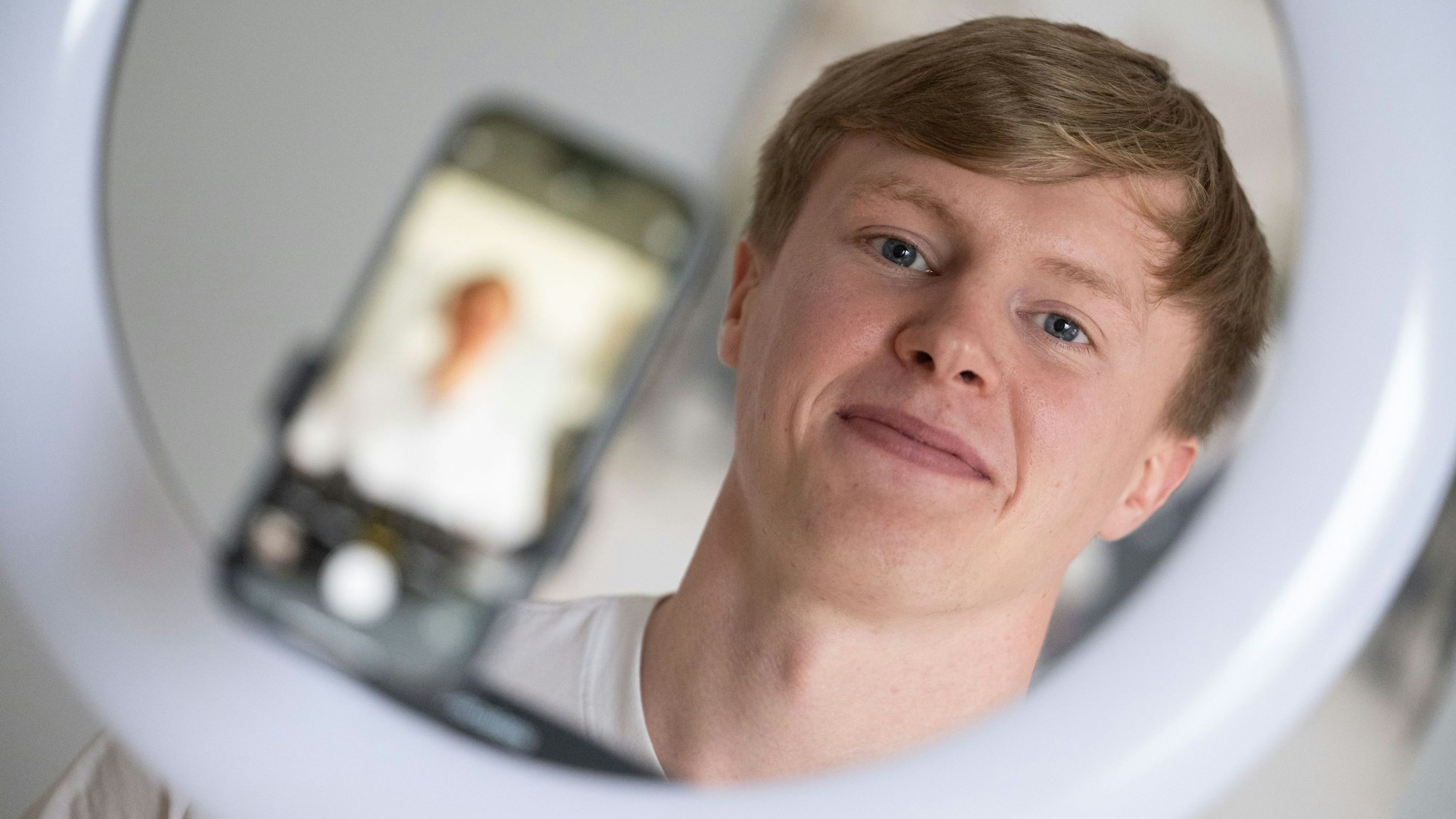 18.01.2024, Baden-Württemberg, Crailsheim: Der Influencer Liam Carpenter steht in seiner Wohnung hinter einem Stativ, auf dem ein Smartphone und ein Ringlicht montiert sind.