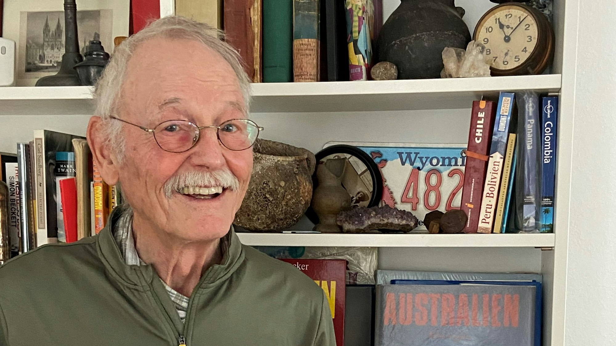 Ein älterer Mann steht vor einem Bücherregal, in dem viele Reisebücher stehen.