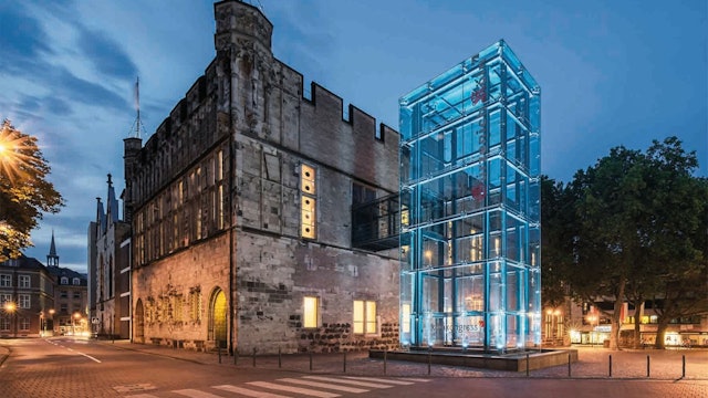 Vor einem Gebäude unter einem dunklen Himmel, der im Begriff ist heller zu werden, steht ein gläserner Kasten auf dem „Koelncongress“ gedruckt ist.