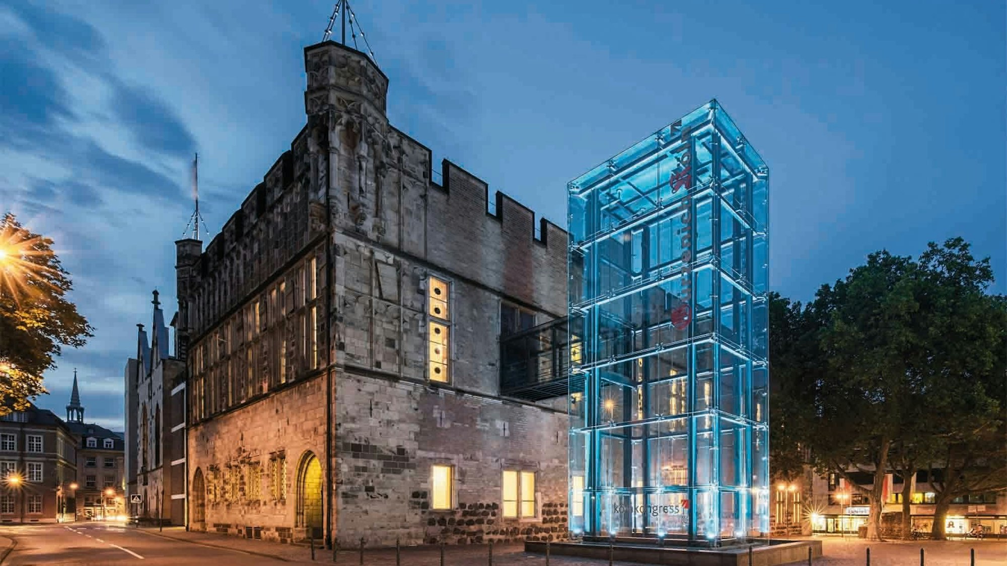 Vor einem Gebäude unter einem dunklen Himmel, der im Begriff ist heller zu werden, steht ein gläserner Kasten auf dem „Koelncongress“ gedruckt ist.