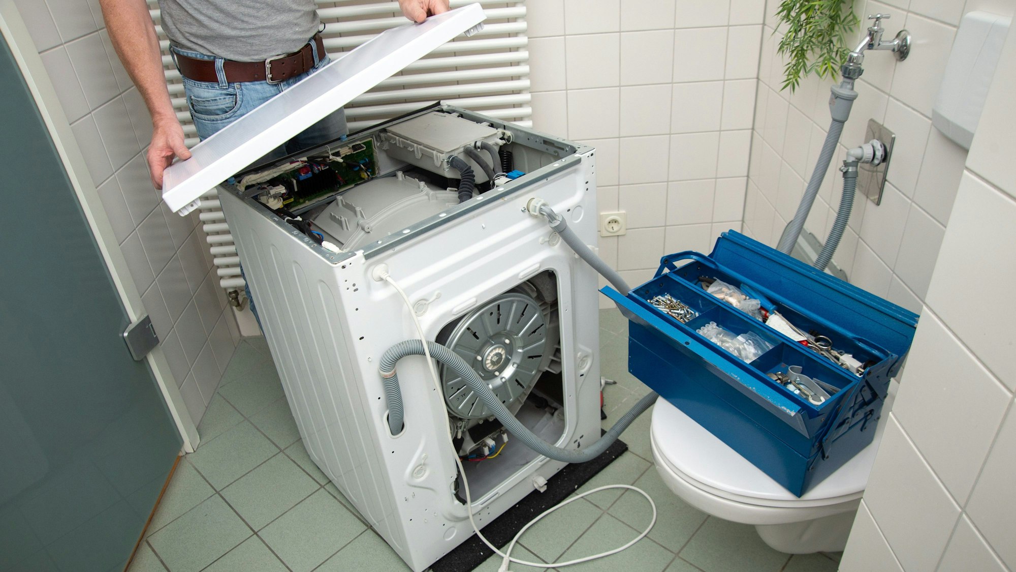 Ein Mann hebt die Abdeckung einer Waschmaschine im Badezimmer hoch. Auf der geschlossenen Toilette steht ein Werkzeugkasten. (Symbolbild)