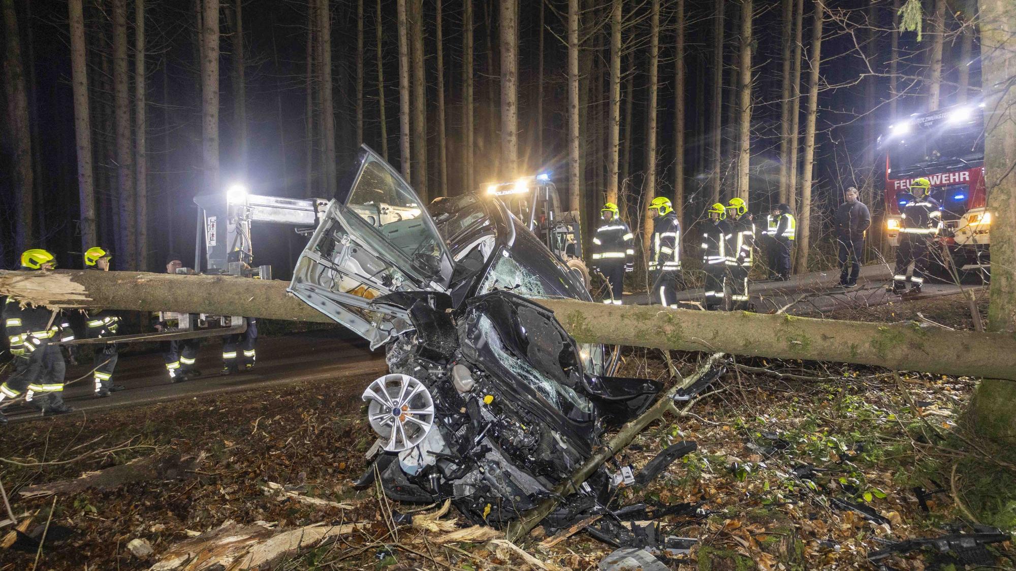 Feuerwehrleute stehen um ein Auto herum, das von einem Baumstamm durchbohrt im Wald liegt.