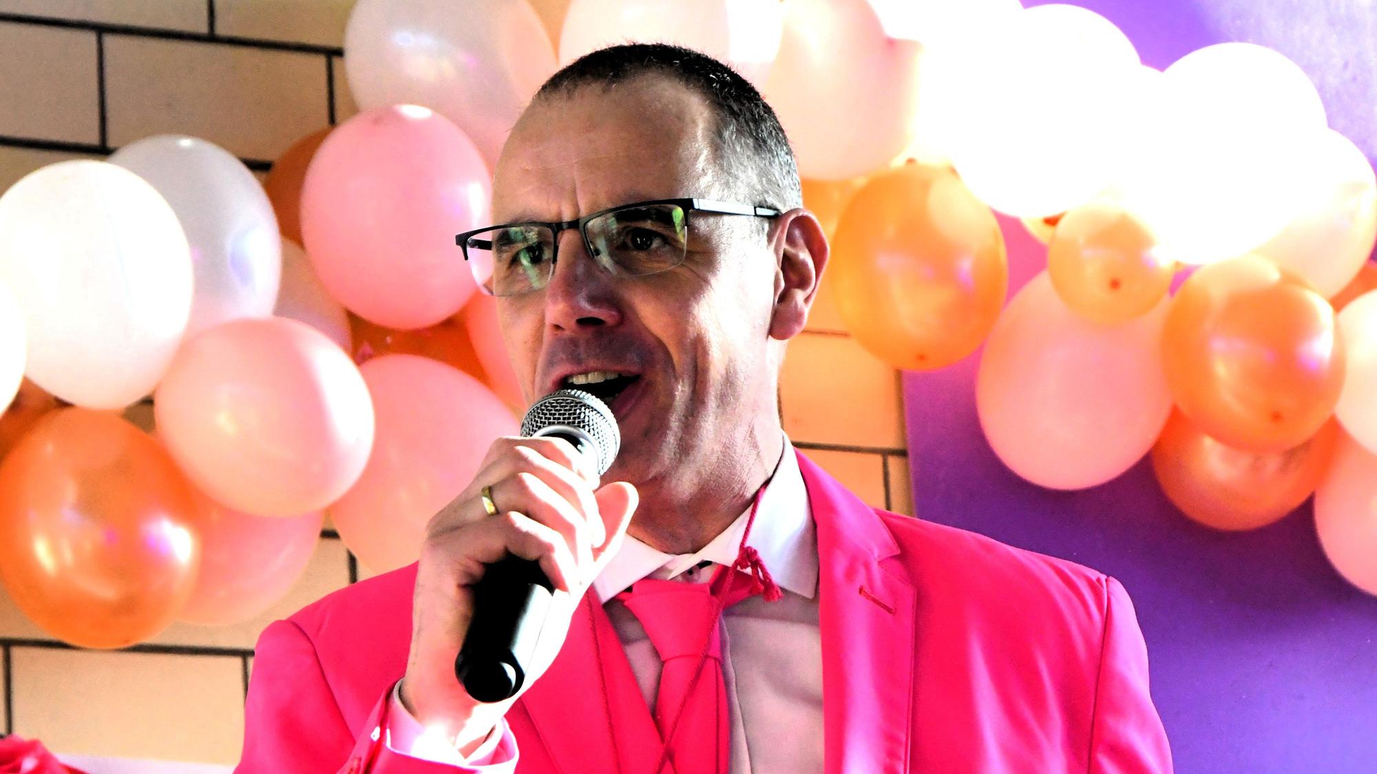 Werner Haag, mit Anzug und Krawatte in Pink, spricht auf der mit Ballons dekorierten Bühne ins Mikrofon.