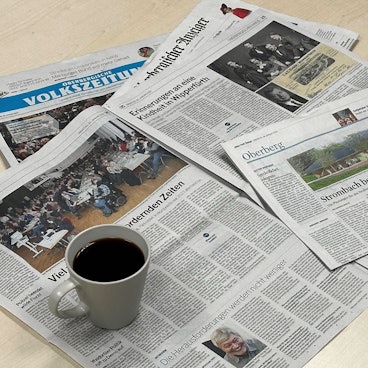 Zeitungsausgaben der Oberbergischen Volkszeitung und des Oberbergischen Anzeigers liegen auf einem Tisch. Darauf steht eine Tasse Kaffee.