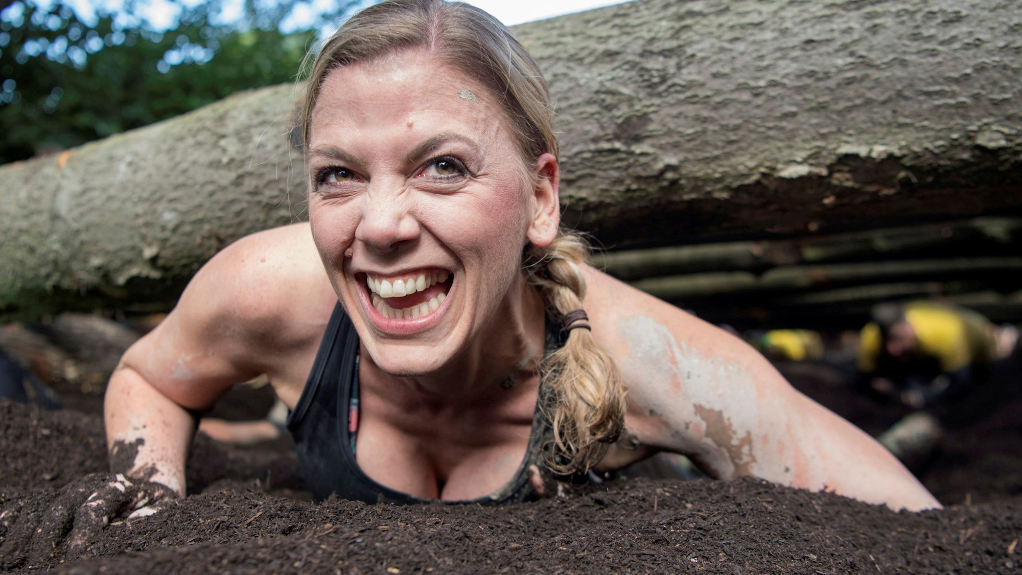 Das Bild zeigt eine Teilnehmerin, die unter Baumstämmen durch robbt und dabei in die Kamera lächelt.