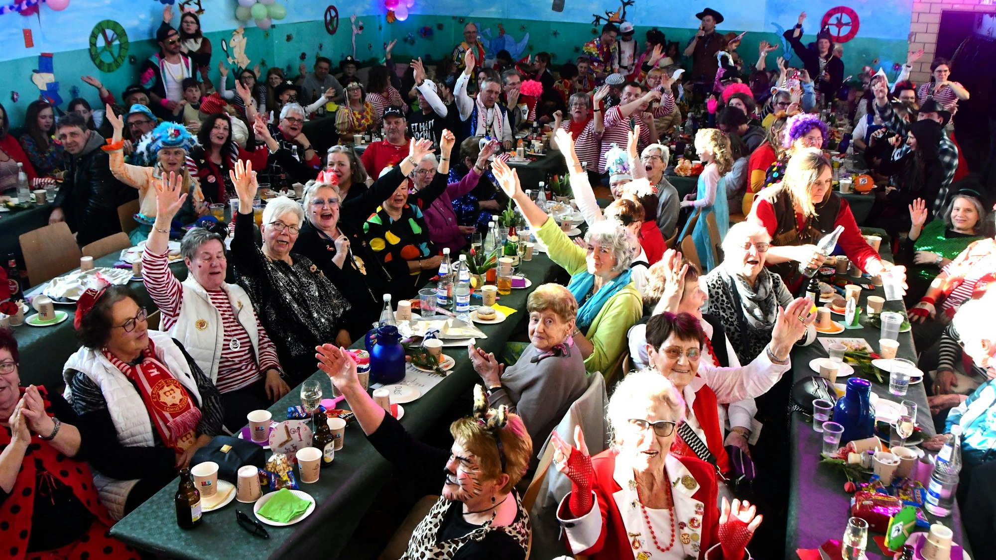 Bunt kostümierte Jecke sitzen in der Dorfhalle Mahlberg eng beisammen an Tischen. Viele lachen und recken zum Alaaf-Ruf die Arme in die Höhe.