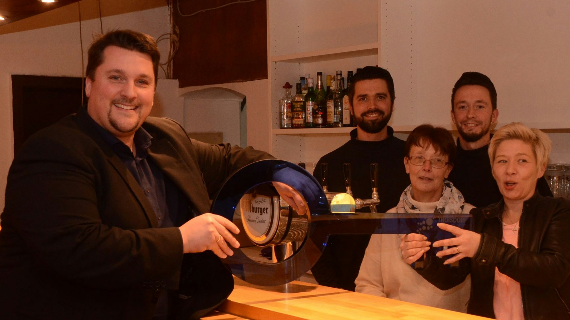 Zwei Frauen und zwei Männer hinter einer Schanktheke. Vor dem Tresen steht ein Mann im dunklen Sakko und Hemd. Sie halten einen Schlüssel aus blauem Plexiglas.